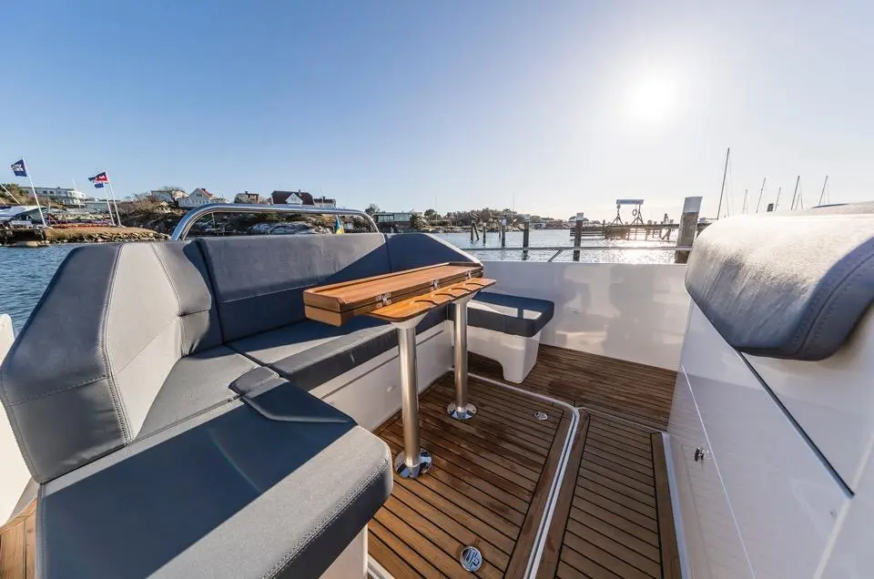 A spacious boat seating area features grey cushions and a wooden table, with a scenic coastal view and soft sunlight.